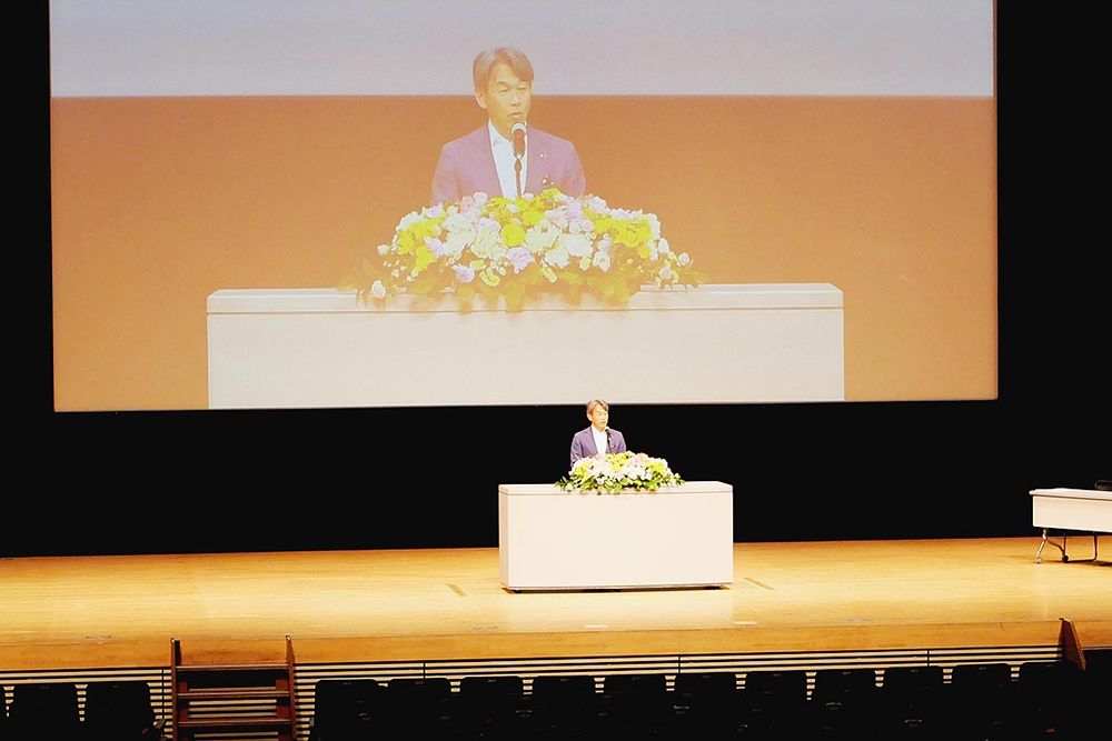 大会冒頭、あいさつに立つ飯塚社長
